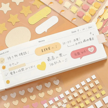 A white notebook or planner page with cute pastel stickers and handwritten notes, surrounded by matching star, heart, and square stickers on a peach-colored surface, showing the possible uses of Daily Palette stickers by BGM Life. 