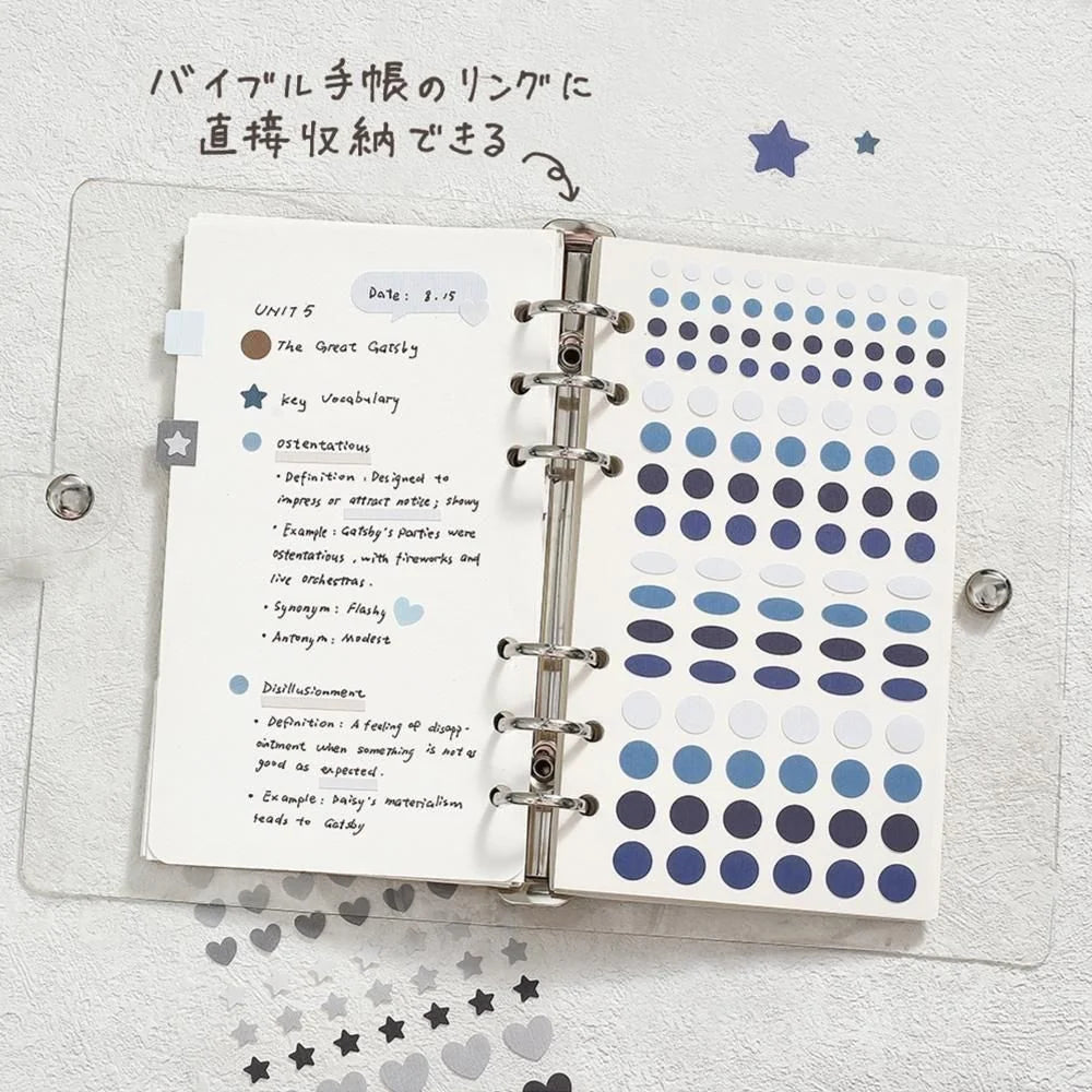 An open clear plastic ring binder notebook on a white textured background, filled with handwritten notes in Japanese and rows of blue and navy circular stickers from the series Daily Palette by BGM Life. Handwritten text at the top says it can store the sticker sheet directly in a ring bound planner or journal.