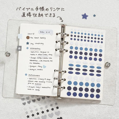 An open clear plastic ring binder notebook on a white textured background, filled with handwritten notes in Japanese and rows of blue and navy circular stickers from the series Daily Palette by BGM Life. Handwritten text at the top says it can store the sticker sheet directly in a ring bound planner or journal.