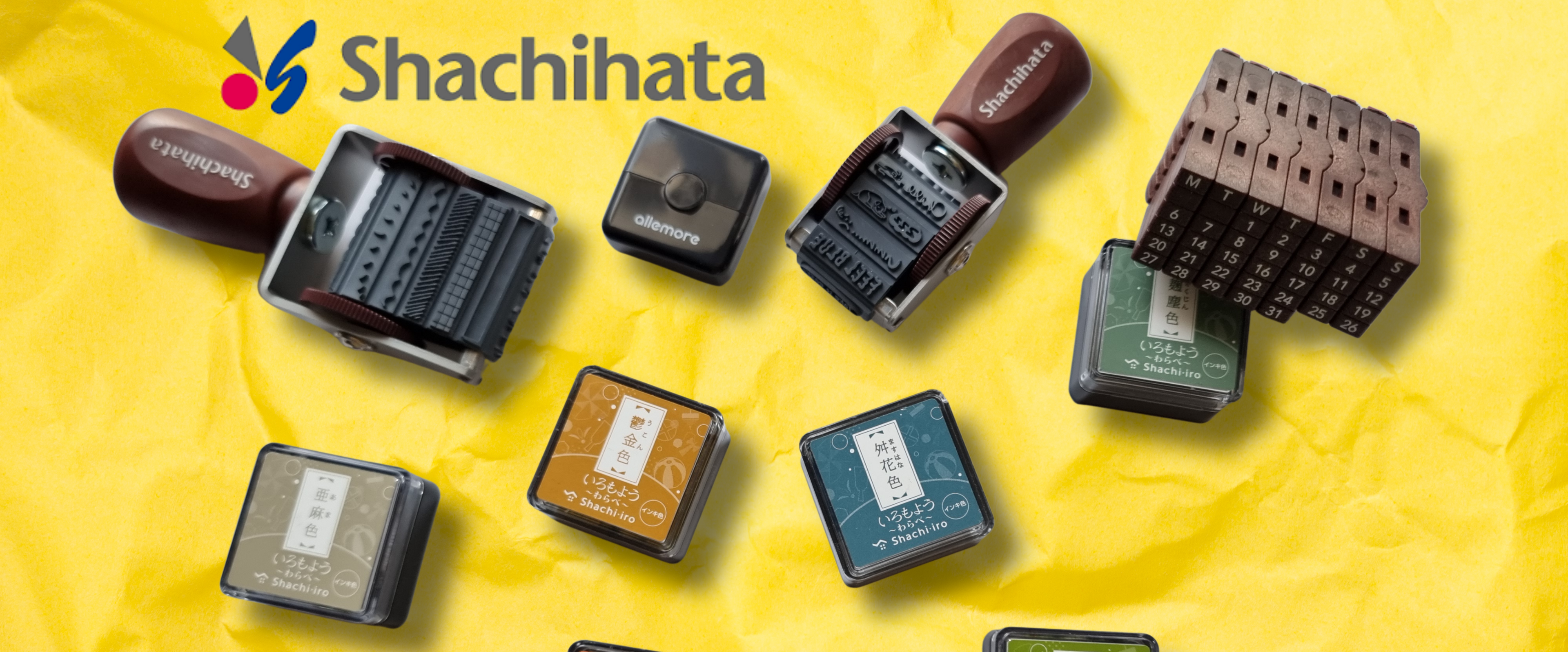 A set of Shachihata rubber stamp and ink products arranged on a bright yellow crumpled paper background. The image shows several stamp handles with dark handles and multiple ink pads in different colors including orange, green, blue, brown, and black.