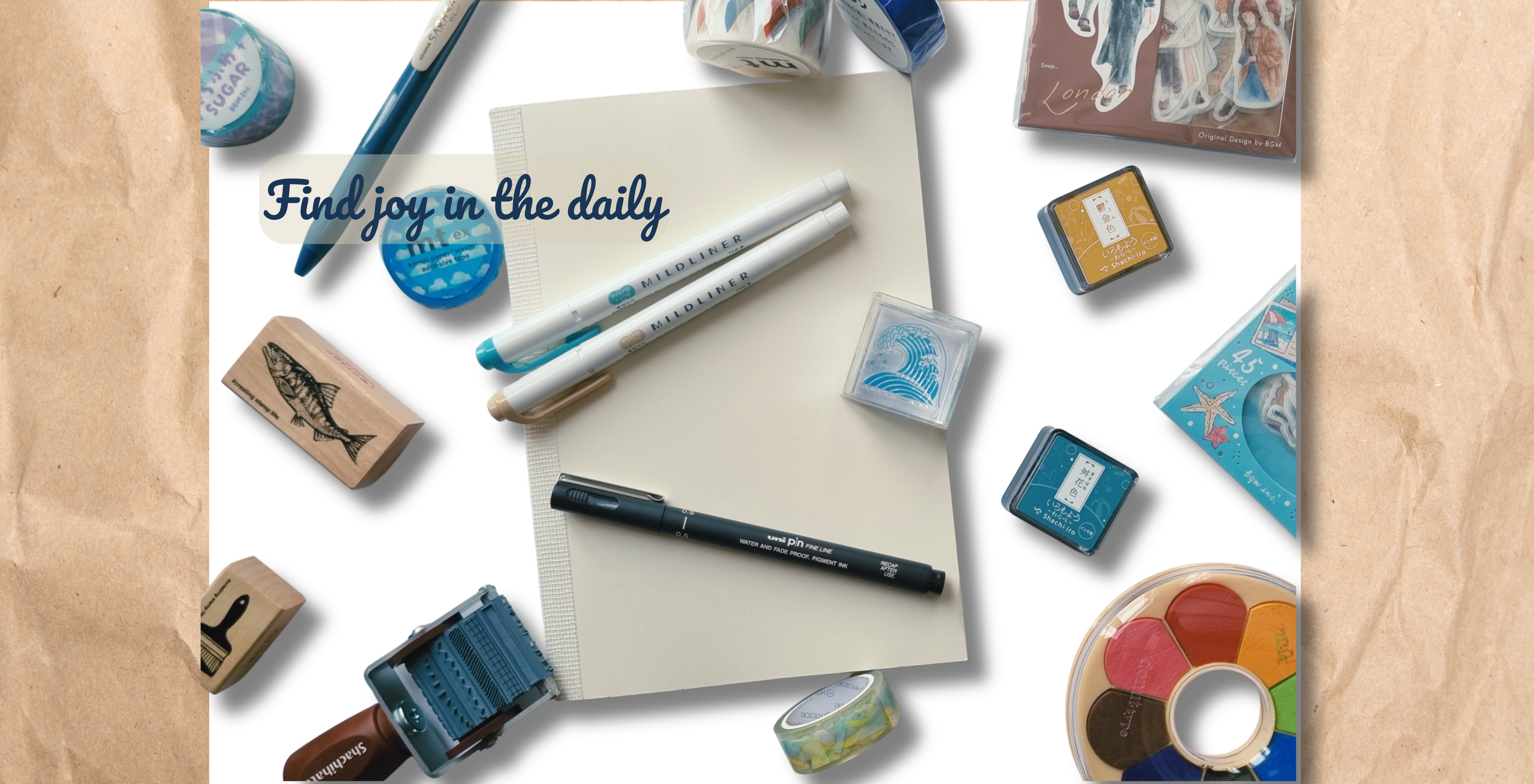 A styled flat-lay of stationery and craft supplies on a white background with brown paper border elements. The central subject is a cream-colored notepad with the text 'Find joy in the daily' written in navy blue script, surrounded by pens, washi tape, rubber stamps, ink pads, and a small paint palette.